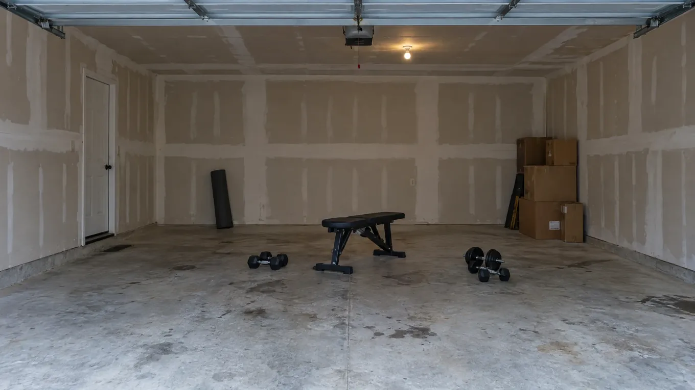 Bare two-car garage with a few loose dumbbells, a bench, unfinished drywall and stained concrete